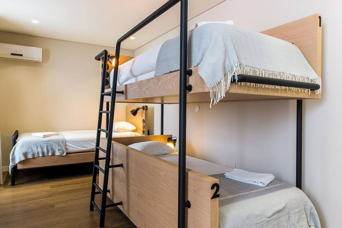Habitació amb lliteres, una escala, un altre llit i un terra de fusta.