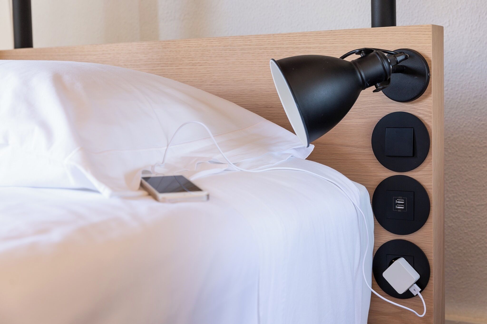 Un lit avec du linge de lit blanc, un téléphone intelligent posé sur le lit, une lampe noire et une tête de lit en bois dotée de trois ports de chargement circulaires.