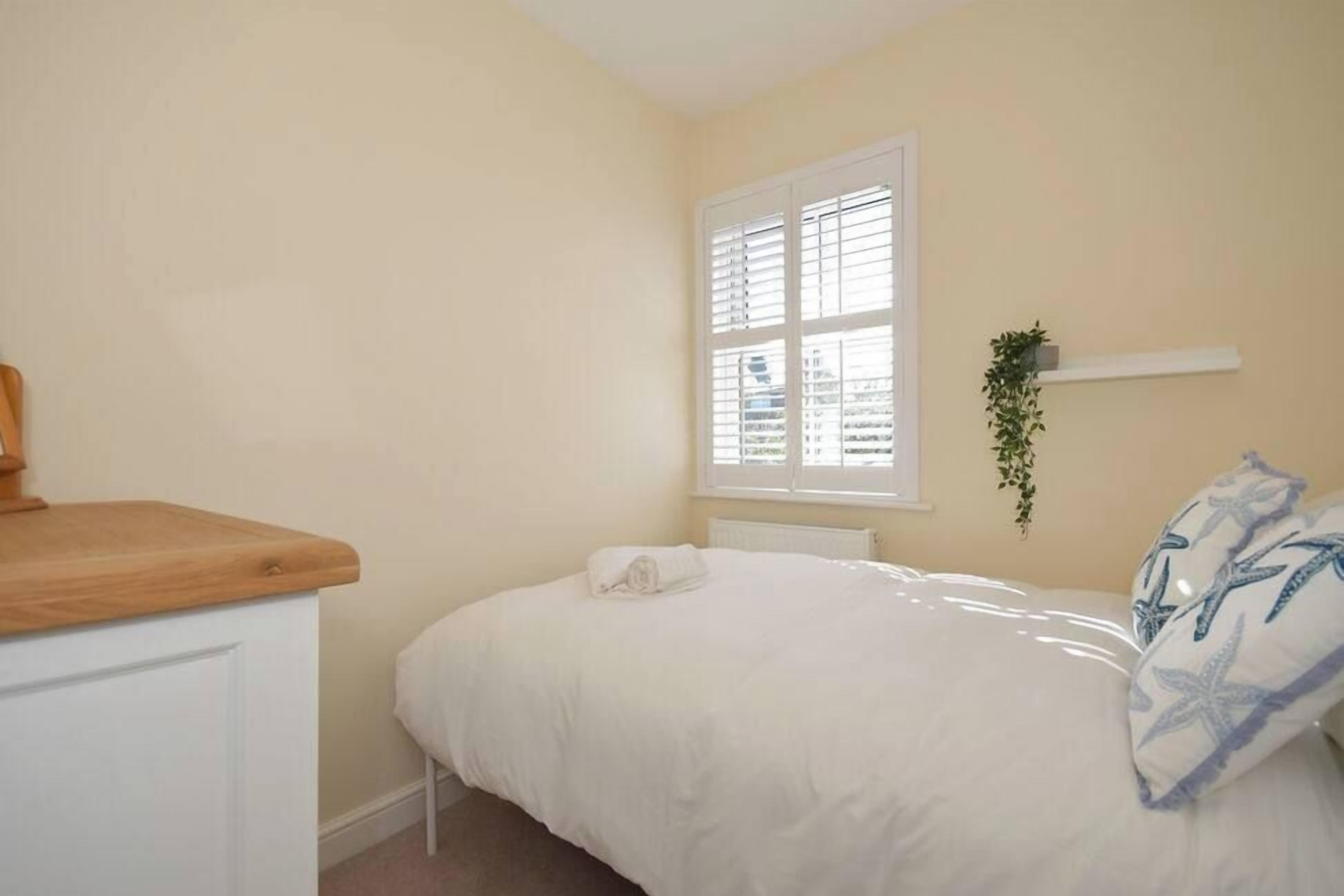 Une chambre avec un lit, une fenêtre avec des stores, un radiateur blanc, un comptoir en bois et une petite plante posée sur une étagère.