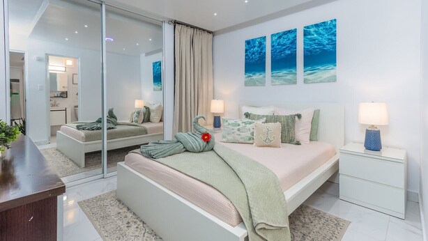 A modern bedroom with a glass partition, a bed with green bedding, two bedside tables with lamps, and three wall-mounted paintings. - Palomino & Icacos Trips / Near El Yunque & Bio Bay (Fajardo)