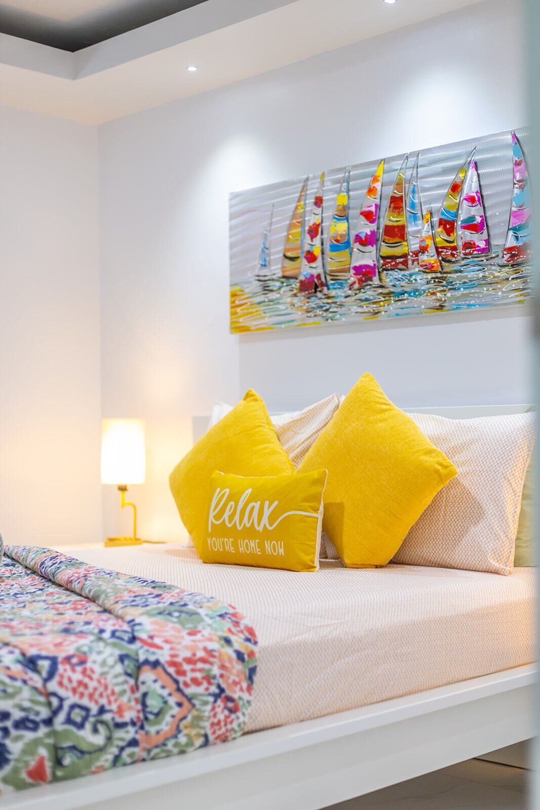 Quarto com cama, almofadas, mesa de cabeceira, abajur e pintura de barcos a vela na parede.