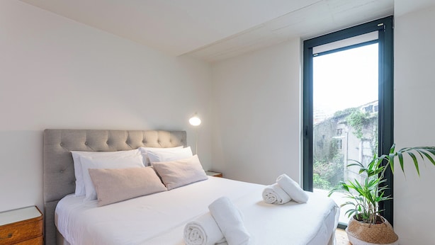 A bedroom with a large bed, a bedside table, a lamp, a window, and a potted plant. - Foz by the River (Porto)
