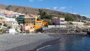 Una località costiera con edifici lungo la riva, una spiaggia e una collina rocciosa sullo sfondo.