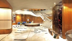Eine geräumige Lobby mit einer geschwungenen Sitzecke, einem zentralen runden Tisch und einer Treppe, die in ein Obergeschoss führt.