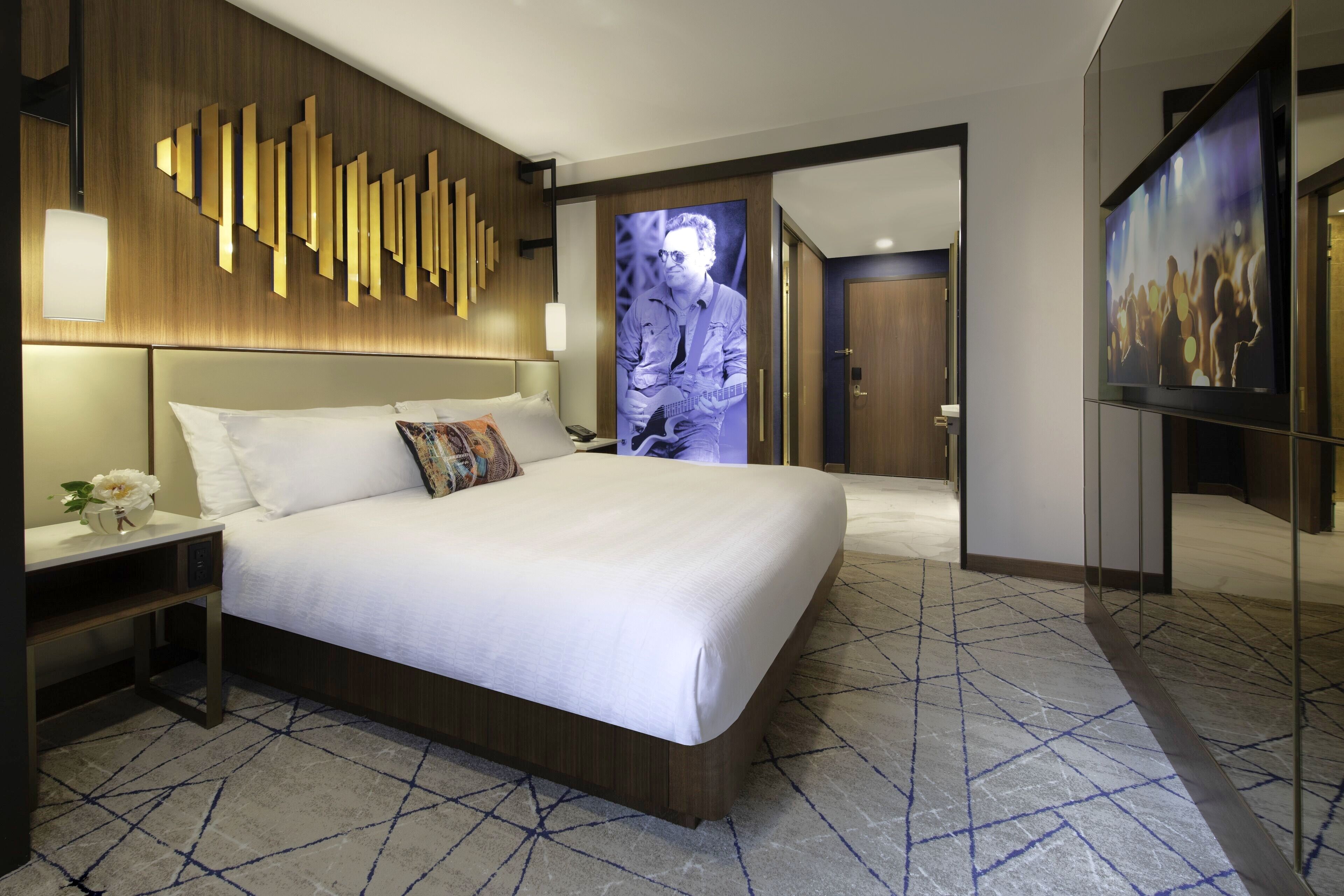 A modern hotel room with a large bed, a wooden headboard, a nightstand, a bedside table with a vase of flowers, and a large wall-mounted TV.
