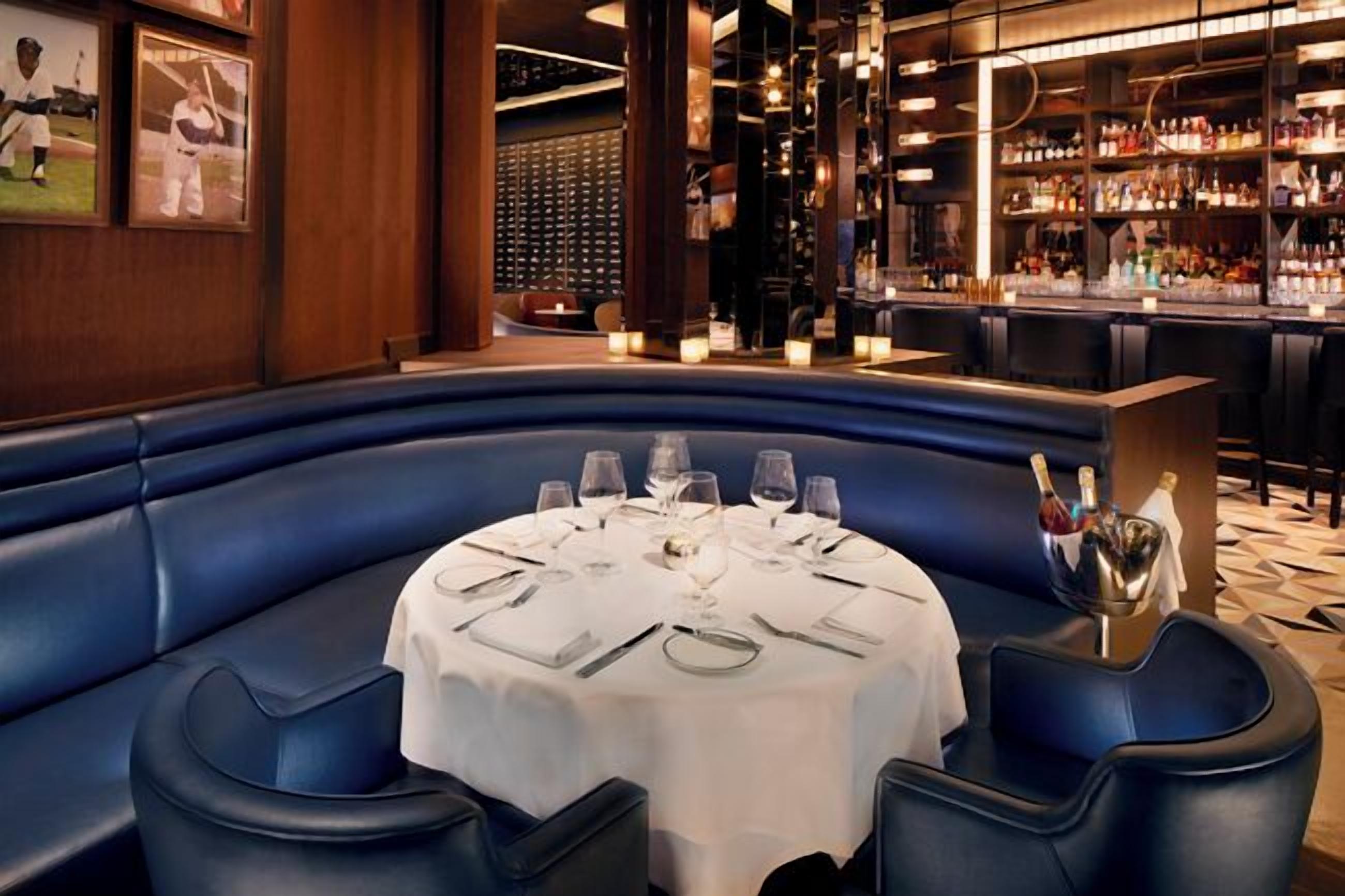 A restaurant with a round table set for dining, blue cushioned seating, and a bar area with shelves of bottles.