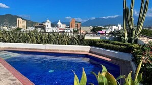 Une piscine à l’eau d’un bleu cristallin, entourée de plantes et de cactus, avec des bâtiments et des montagnes en arrière-plan.