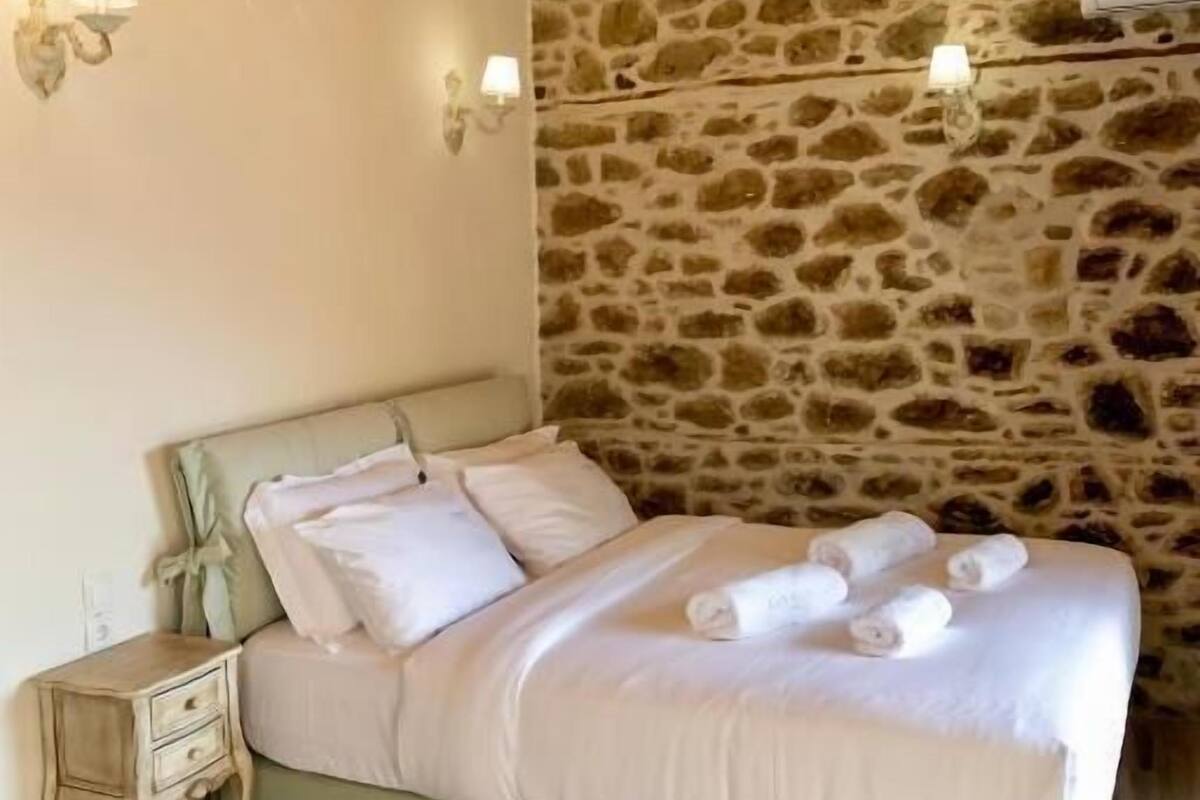 A bed with white linens and rolled towels, a wooden nightstand with a drawer, a stone wall, and a wooden ceiling.