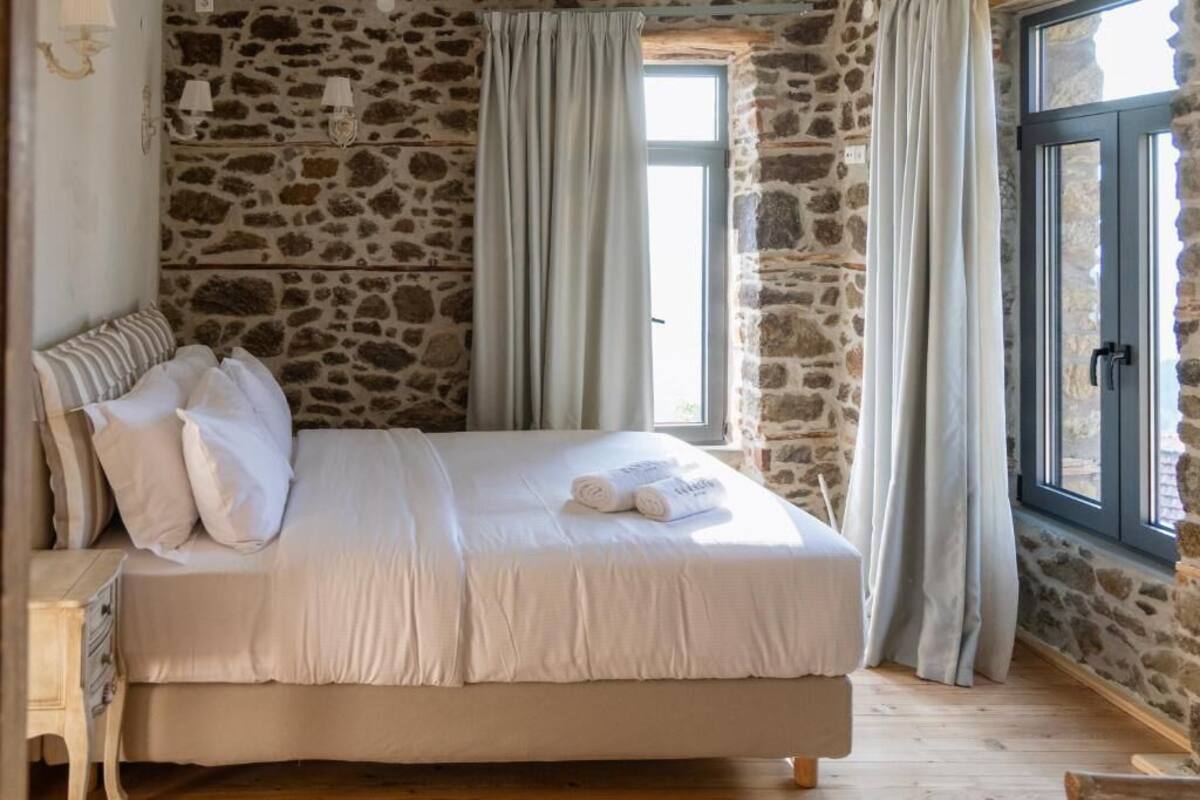 A bedroom with a stone wall, a bed with white bedding, a nightstand, and a window with curtains.