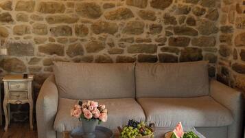 Interior de pared de piedra con un sofá, una mesa con flores y fruta, y una mesita de noche con una lámpara.