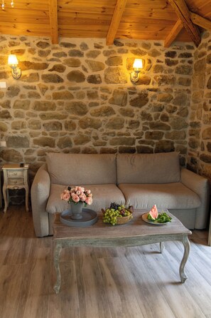 Interior de pared de piedra con un sofá, una mesa con flores y fruta, y una mesita de noche con una lámpara.