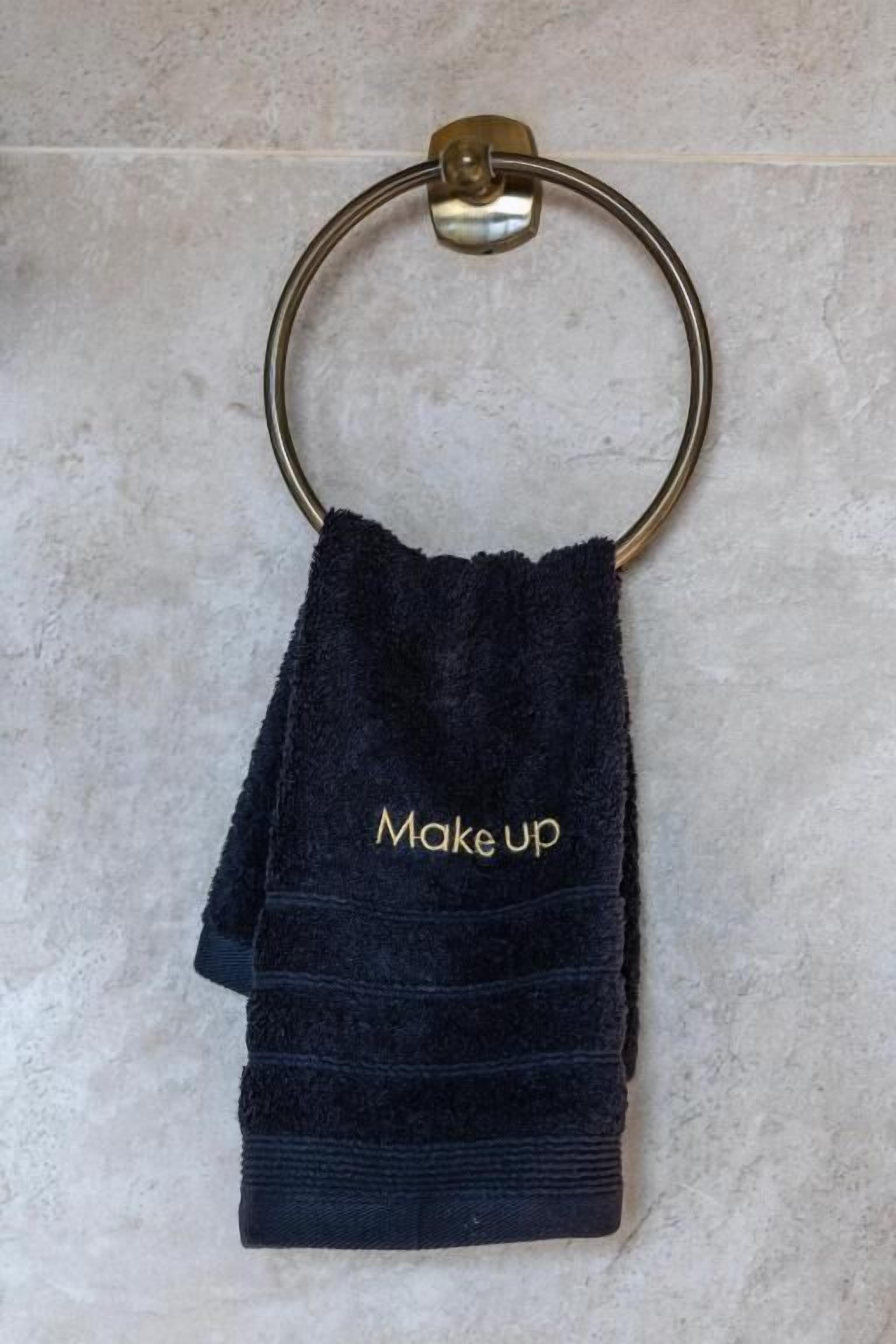 Et mørkeblåt håndklæde med 'Make up' broderet i guld, hængende på en messinghåndklædering.