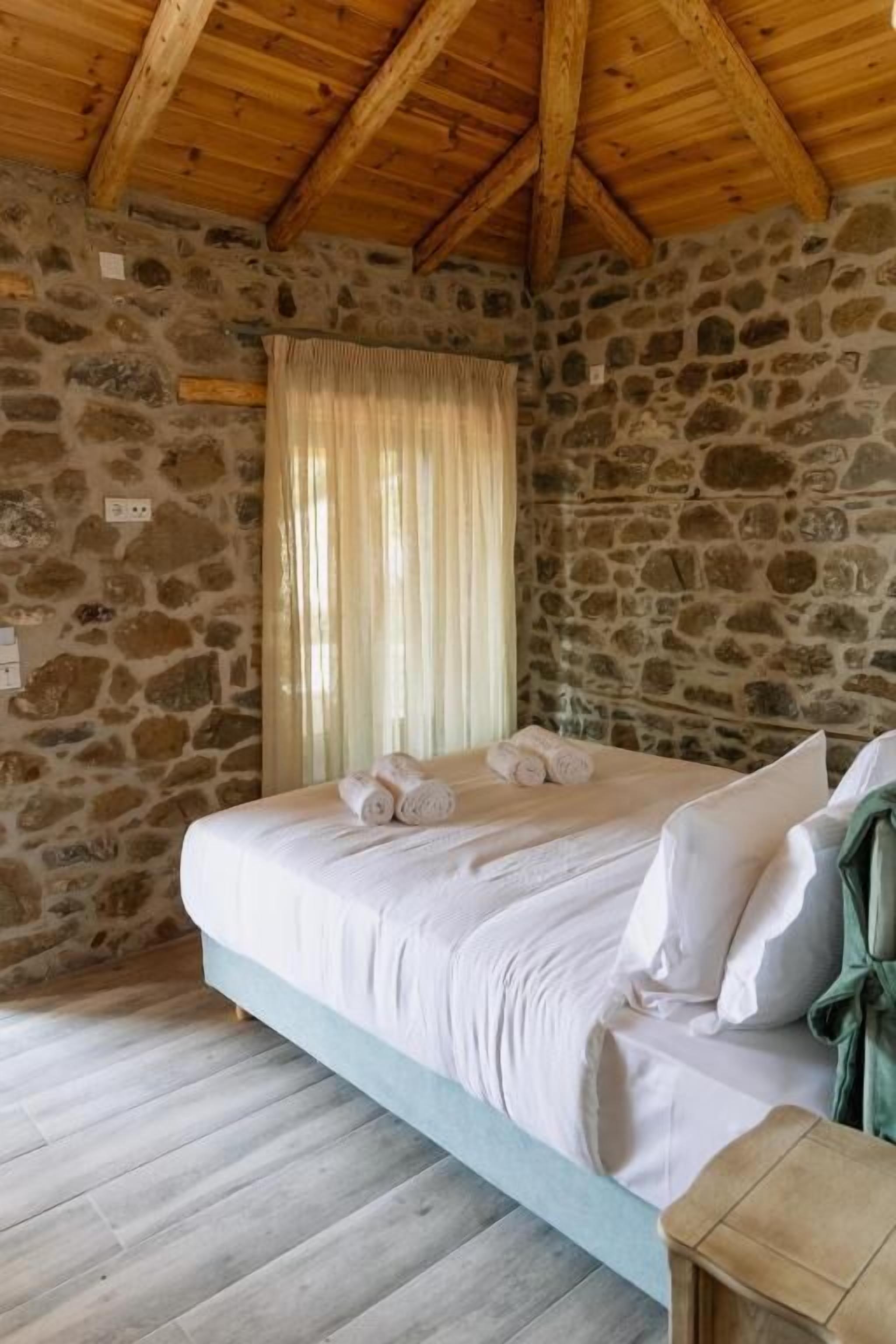 A bedroom with a stone wall, wooden ceiling beams, a bed with white linens, and a green towel on the side.