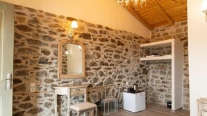 Um quarto com paredes de pedra, teto de madeira, um espelho, uma mesa pequena, um banco, um armário e um cabideiro.