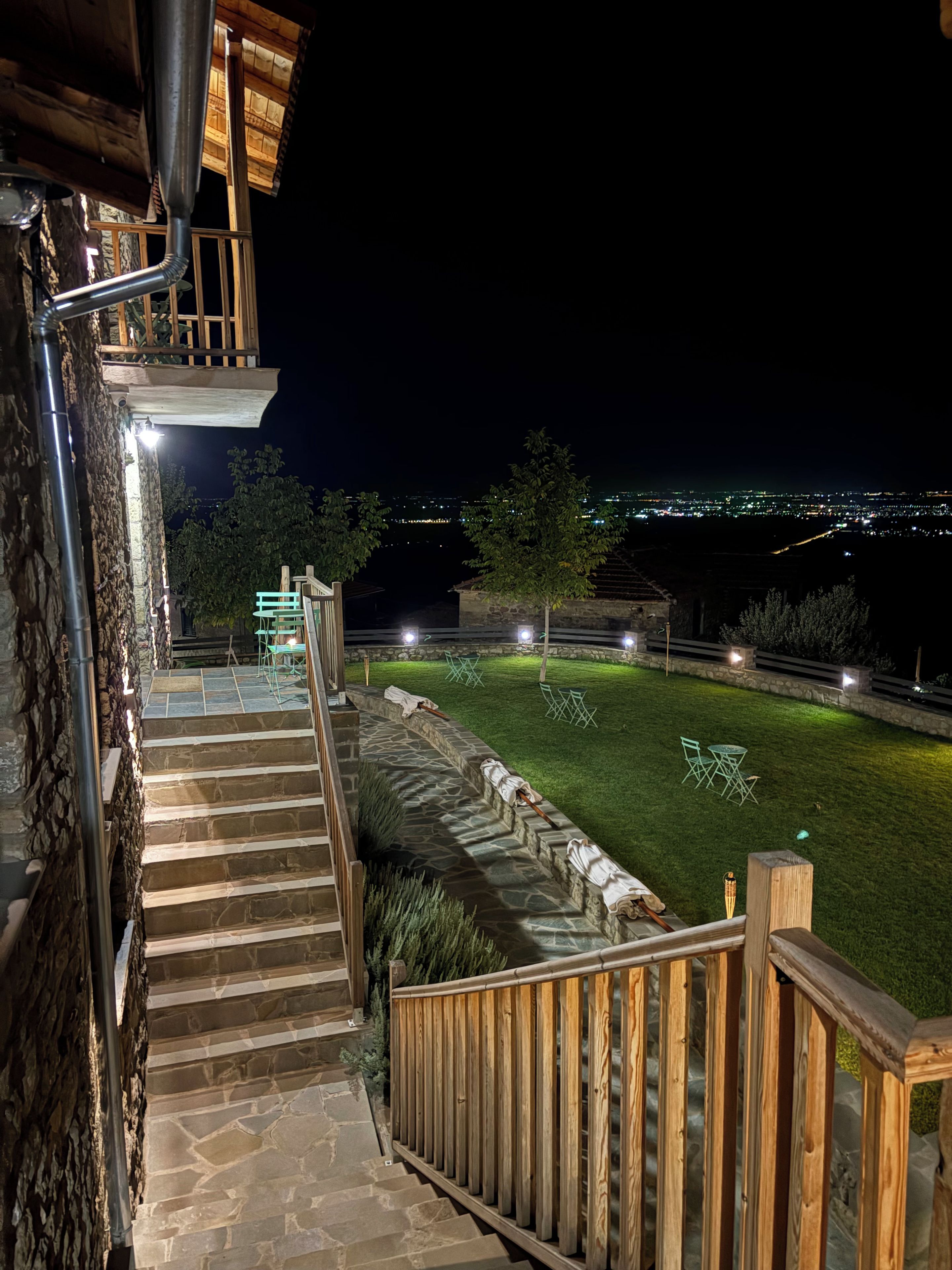 Eine Steintreppe führt zu einem Holzbalkon mit Blick auf eine beleuchtete Stadtlandschaft bei Nacht.