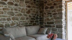 Quarto com parede de pedra, vigas de madeira no teto, sofá cinza e uma pequena mesa com um vaso de flores.