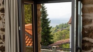 Quarto com parede de pedra, janela com porta deslizante aberta e vista para paisagem verde e edifícios ao longe.