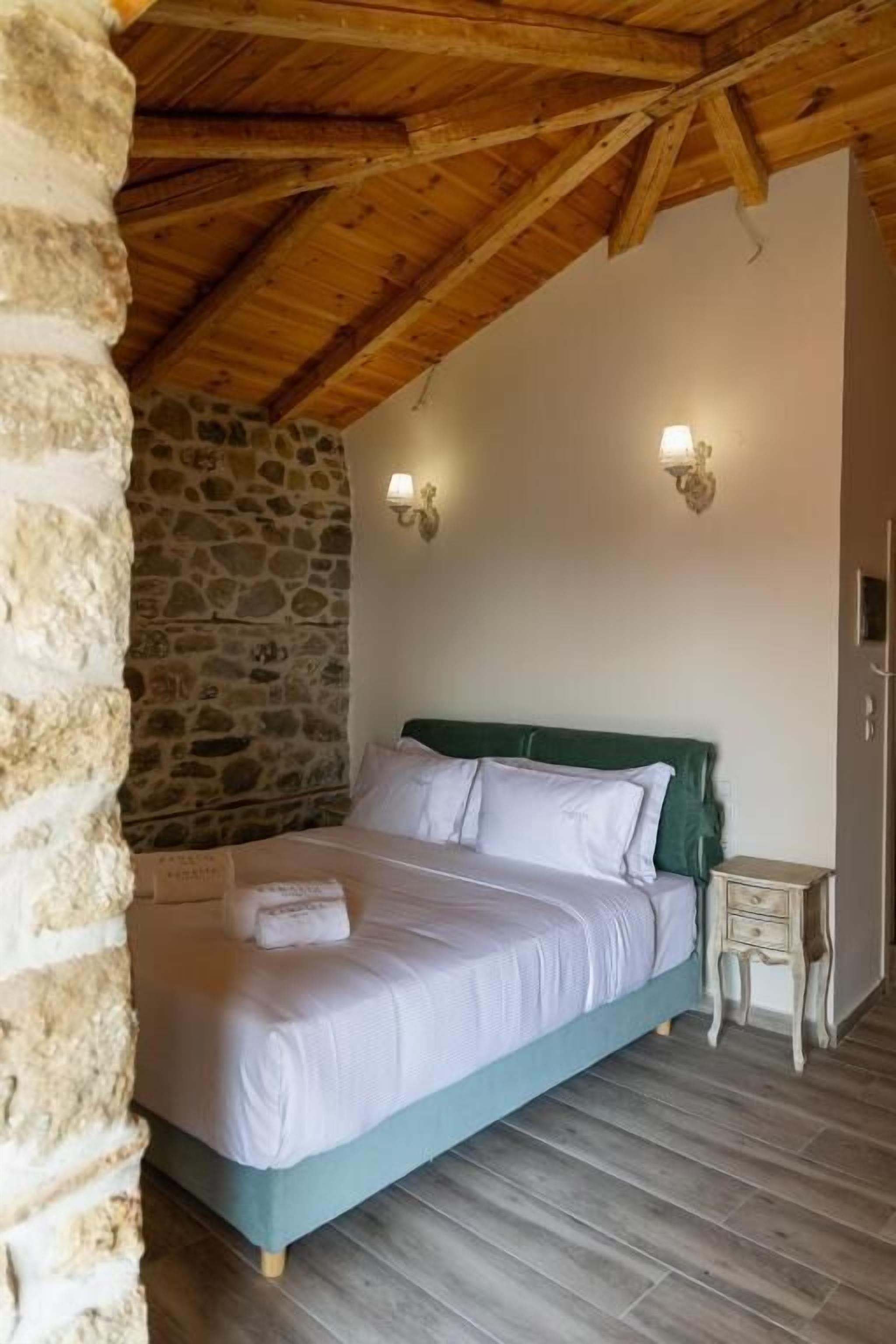 Un dormitorio con pared de piedra, vigas de madera en el techo, cama con ropa blanca y dos lámparas fijadas a la pared.
