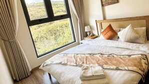 A hotel room with a large window, a bed with white linens, a wooden headboard, and a nightstand with a lamp. - 1991 Home Sapa | Fully Equipped Apartment | 5 min to Sapa Center (Sa Pa)