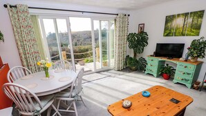 Een woonkamer met een eettafel, stoelen, een houten salontafel, een televisie en een groot raam met gordijnen.