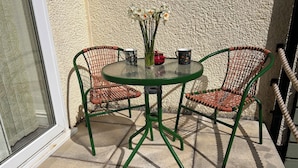 Een groene metalen tafel met twee rieten stoelen, een vaas met witte bloemen en twee gemêleerde mokken tegen een structuurrijke muur.