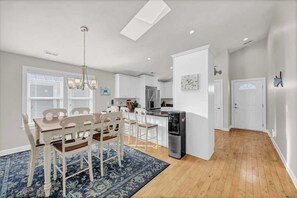 A modern kitchen with white cabinets, a refrigerator, and a dining area with a blue rug and wooden chairs. - Aqua Sea, a beachfront retreat (Carolina Beach)