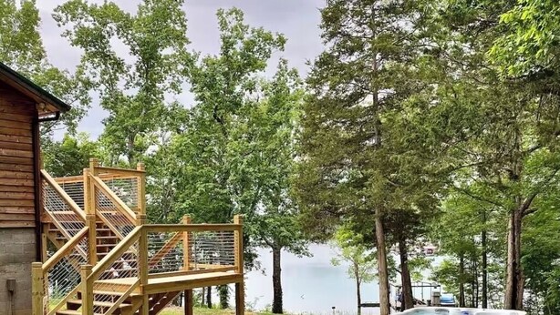 A wooden deck with a staircase leading to a hot tub, surrounded by trees and a body of water. - Stay@Black Sheep:Lakefront•HotTub•No Steps2Dock! Spacious_Location_Dog friendly (New Tazewell)