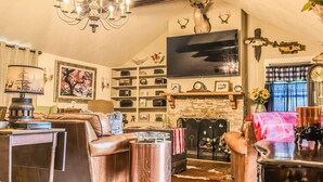 A cozy living room with a fireplace, leather furniture, a chandelier, and a television mounted on the wall. - Almeneiro Cabin - A Charming Nature Retreat (Greensboro)