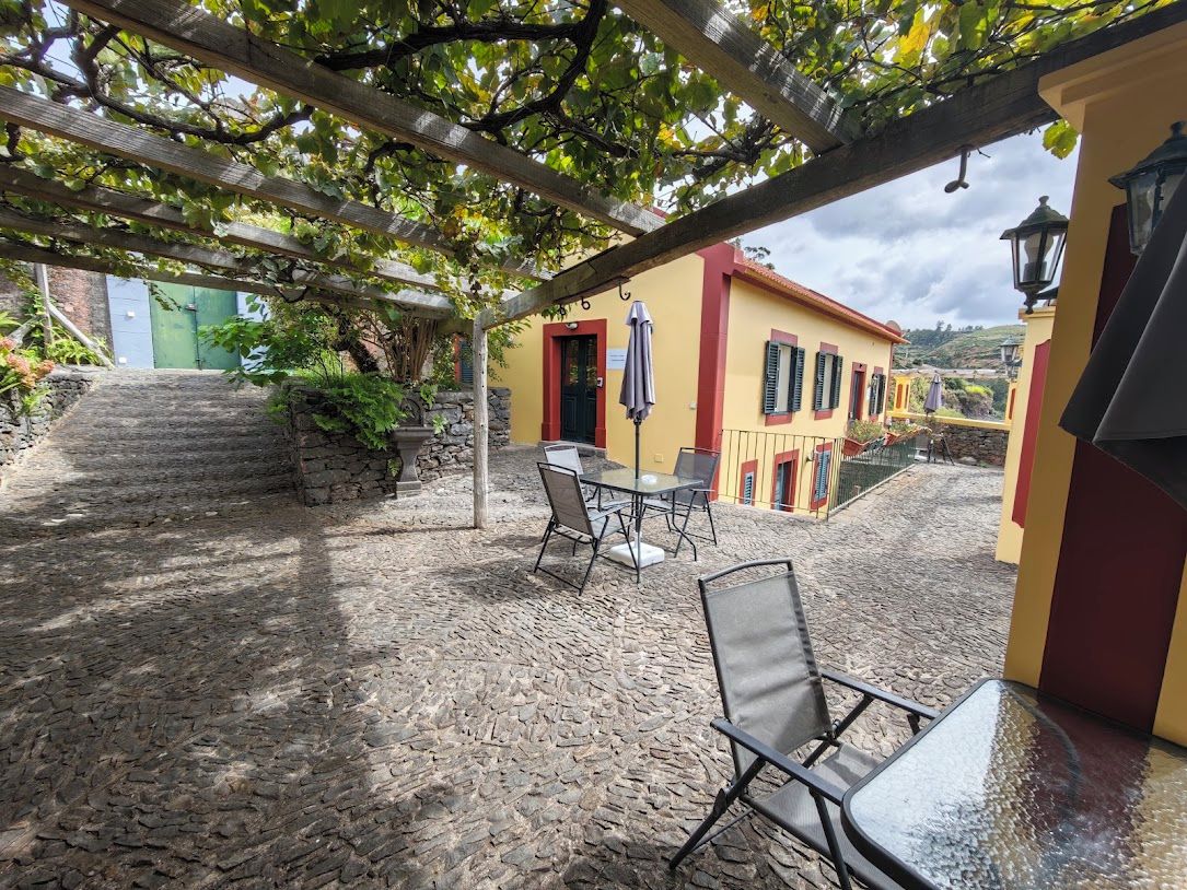 Un patio exterior con piso de piedra, una pérgola con plantas trepadoras y un edificio amarillo con puerta roja.