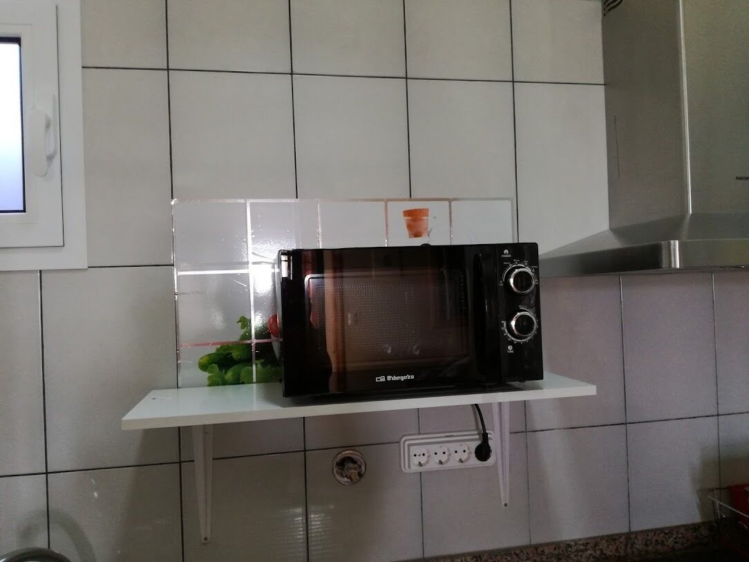 Een keuken met een magnetron op een wit schap, betegelde wanden en een raam.