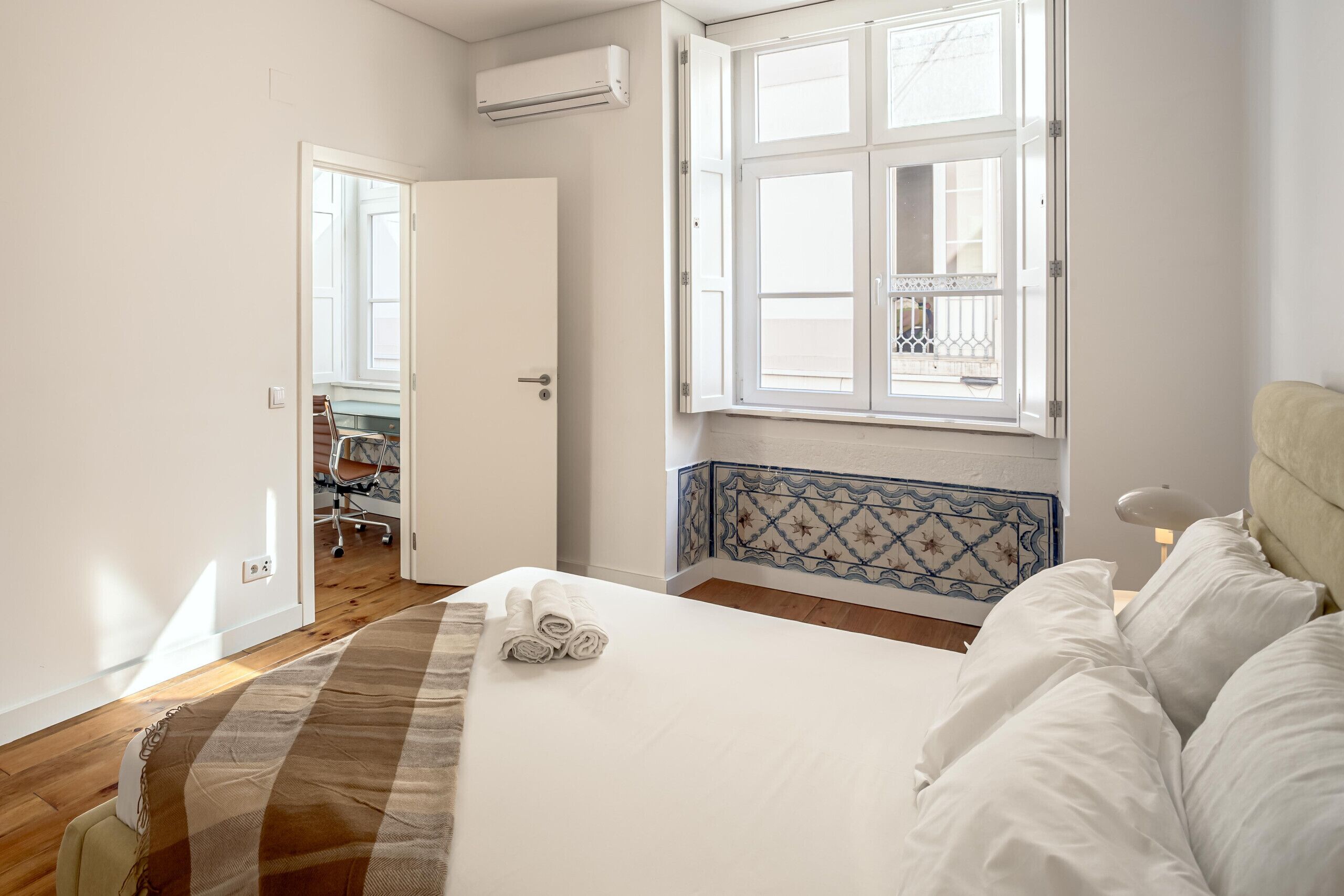 A bedroom with a bed, a window with a view, a door leading to another room, and a wall-mounted air conditioner.