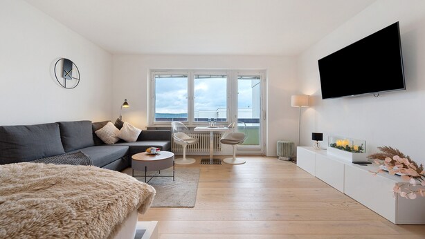 A modern living room with a sofa, a dining table with chairs, a flat-screen TV mounted on the wall, and a large window.