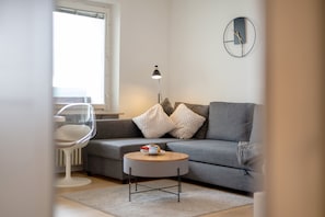 Una sala de estar moderna con un sofá gris, una mesa redonda de centro, una silla transparente, una lámpara de pie y un reloj de pared.