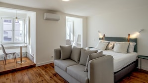 Ein modernes Schlafzimmer mit einem grauen Sofa, einem kleinen runden Tisch und einem Bett mit dunkelgrüner Kopfteil.