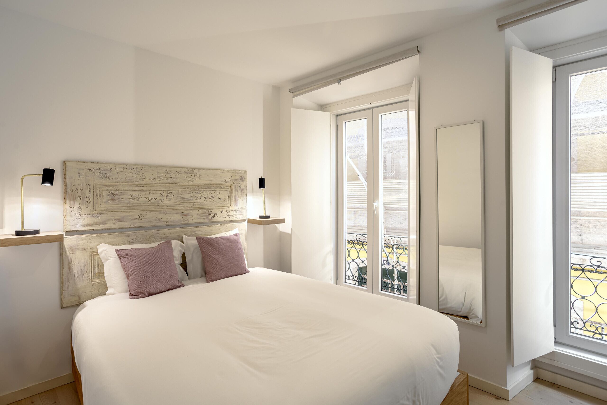 A bedroom with a large bed, a wooden headboard, a window with a view of a balcony, and a mirror on the wall.