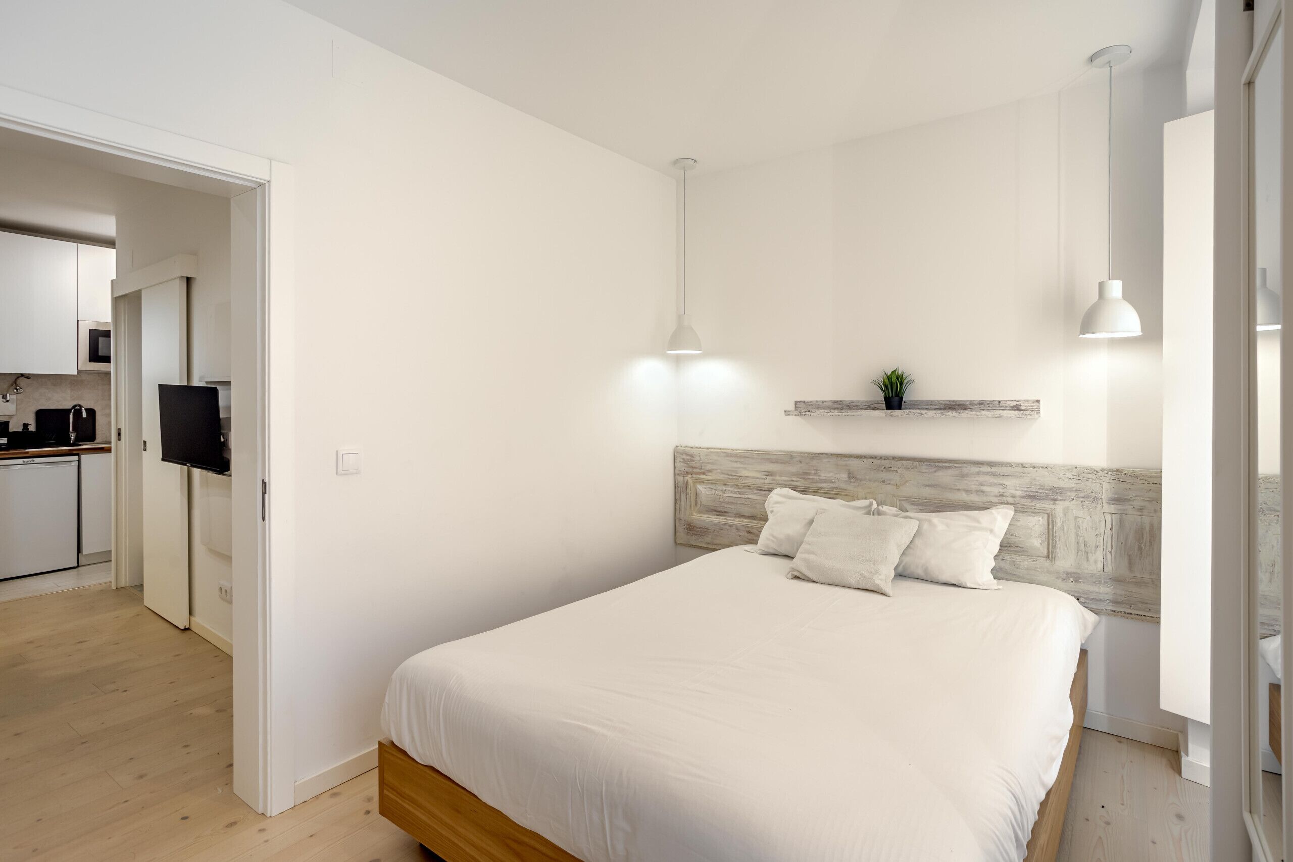 A bedroom with a wooden headboard, a bed with white bedding, a small plant on a shelf, and a TV in the adjacent room.