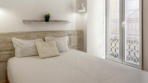 A bedroom with a wooden headboard, a bed with white bedding, a small plant on a shelf, and a balcony with a metal railing. - Downtown Modern Apartments I by Homing (Lisbon)