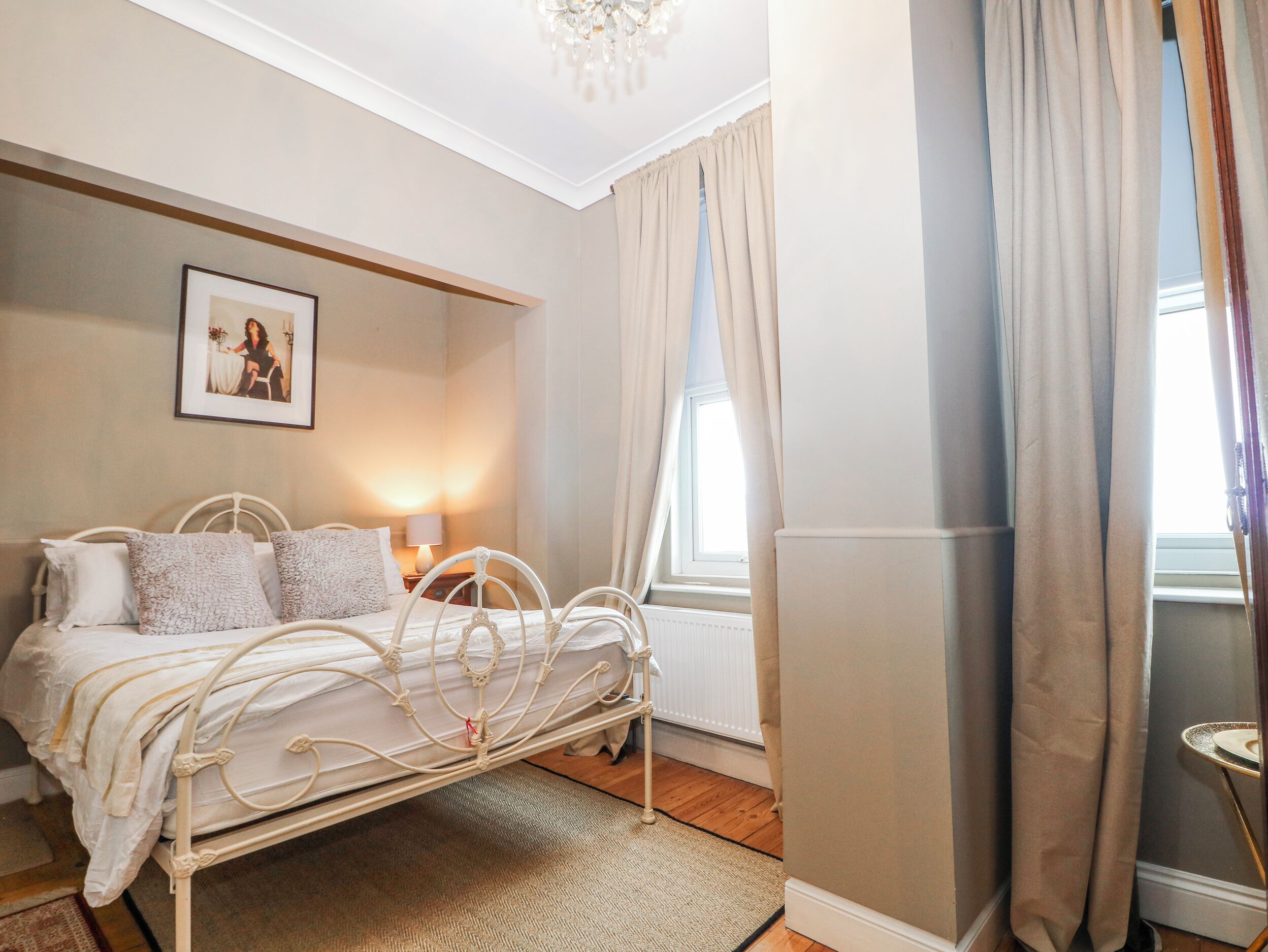 Une chambre avec un lit en métal blanc, une table de chevet avec une lampe, un tableau encadré au mur et une fenêtre avec des rideaux.