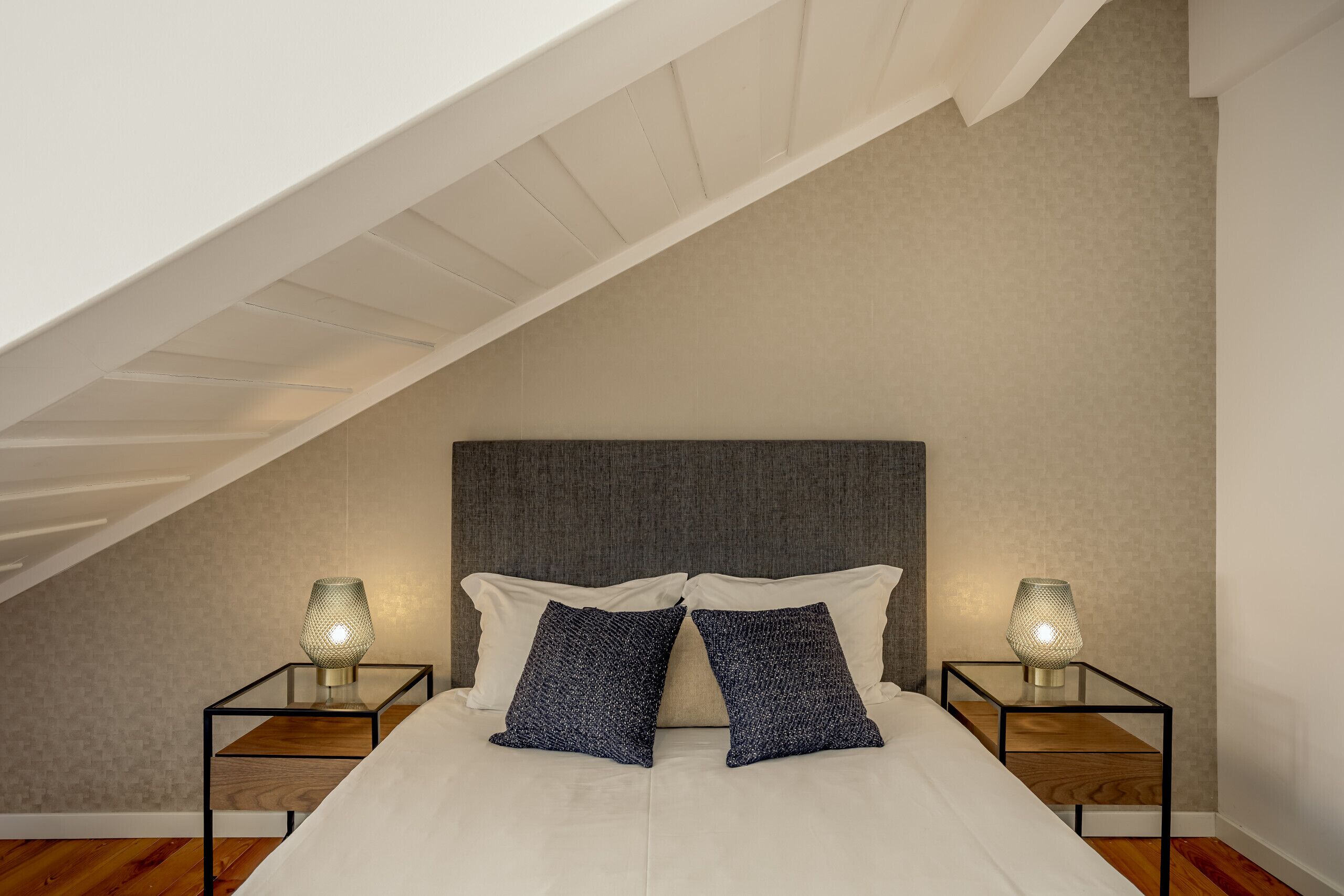A bedroom with a sloped ceiling, a bed with white bedding, two nightstands with lamps, and a dark headboard.