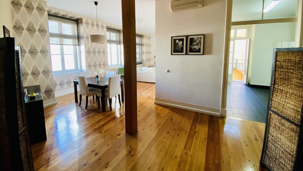 A spacious living area with wooden flooring, a dining table with chairs, a large window, and an air conditioning unit on the wall. - Downtown Riverside III by Homing (Lisbon)