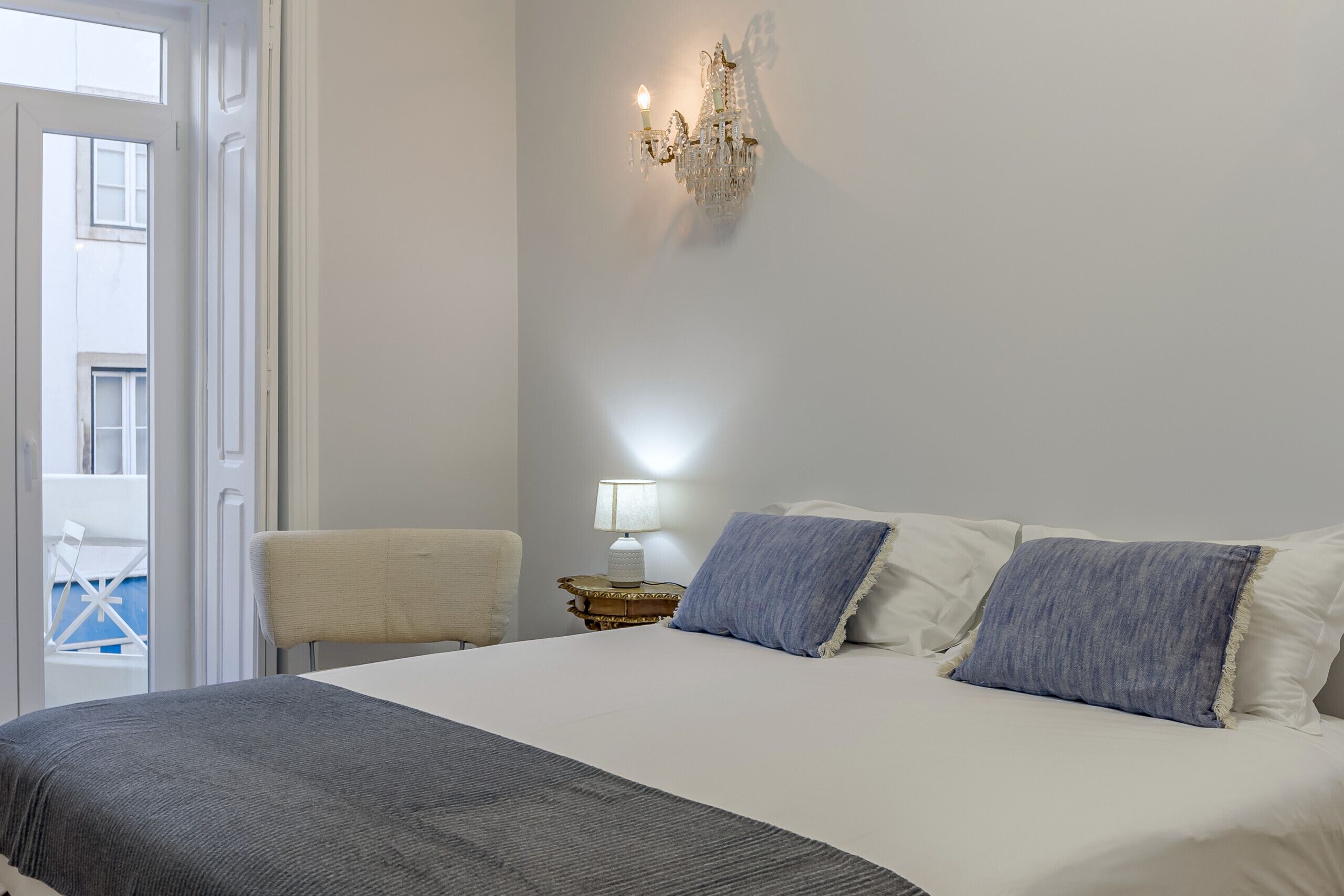 A bedroom with a bed, a chair, a nightstand with a lamp, and a wall-mounted light fixture.