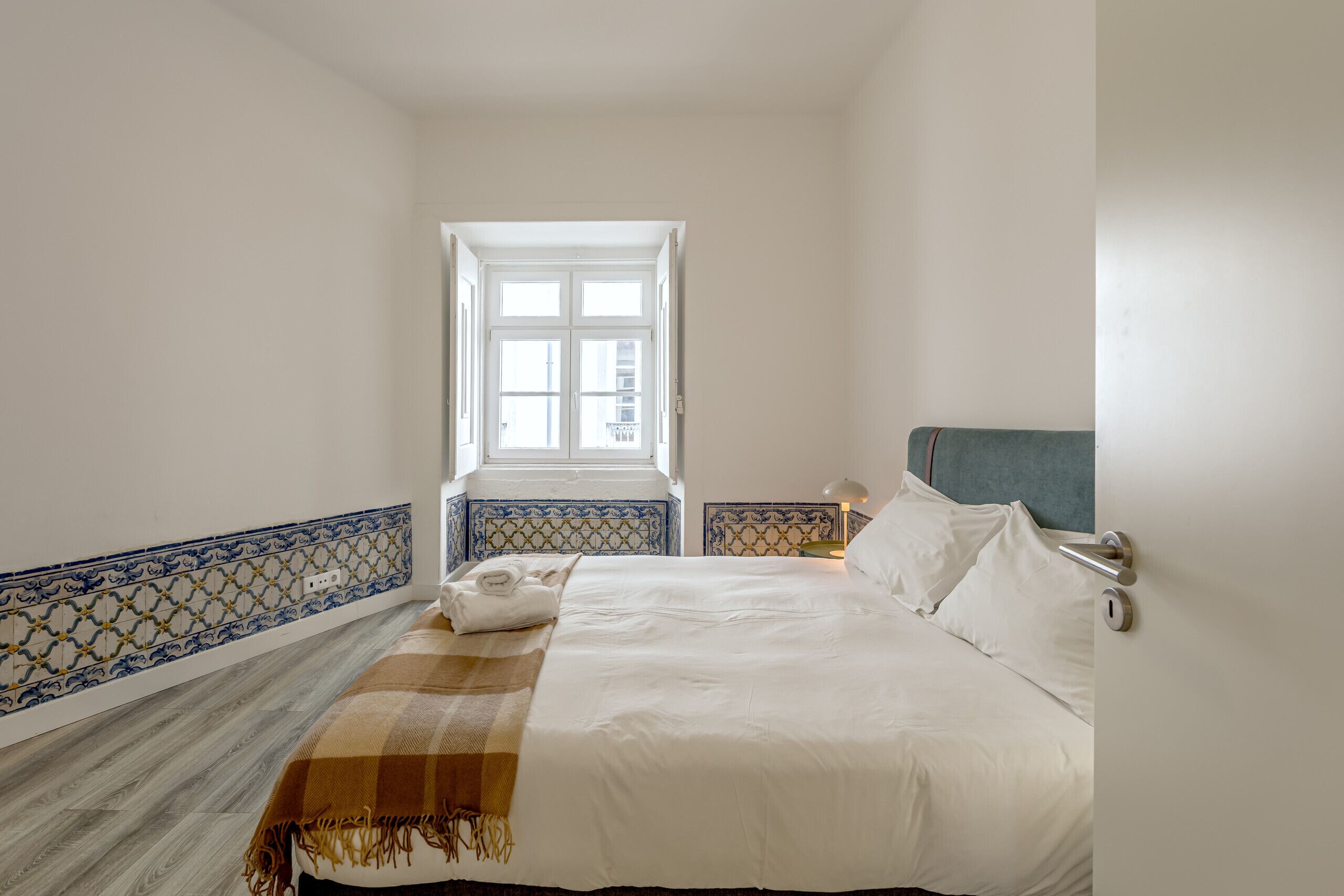 A bedroom with a bed, a window, a nightstand, and a wall with blue and white tiles.