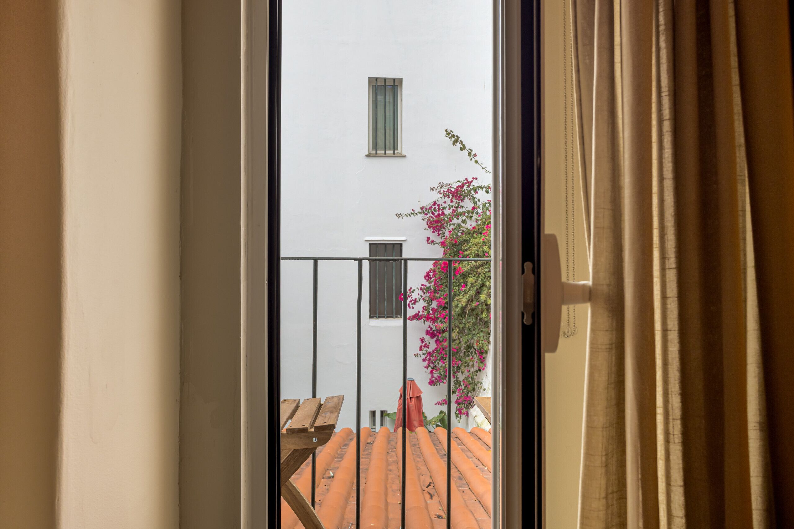Vista attraverso una porta finestra su un edificio con tetto in tegole, una piccola finestra e una pianta in vaso con fiori rosa.