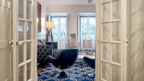 Una habitación con piso estampado, un sillón tapizado oscuro, una lámpara y vistas por una puerta.