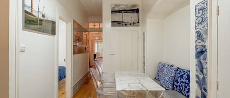 Un pasillo con una zona de comedor que incluye una mesa con cubierta de mármol y sillas transparentes, un sofá con cojines de diseño azul, y una pared con una fotografía en blanco y negro.