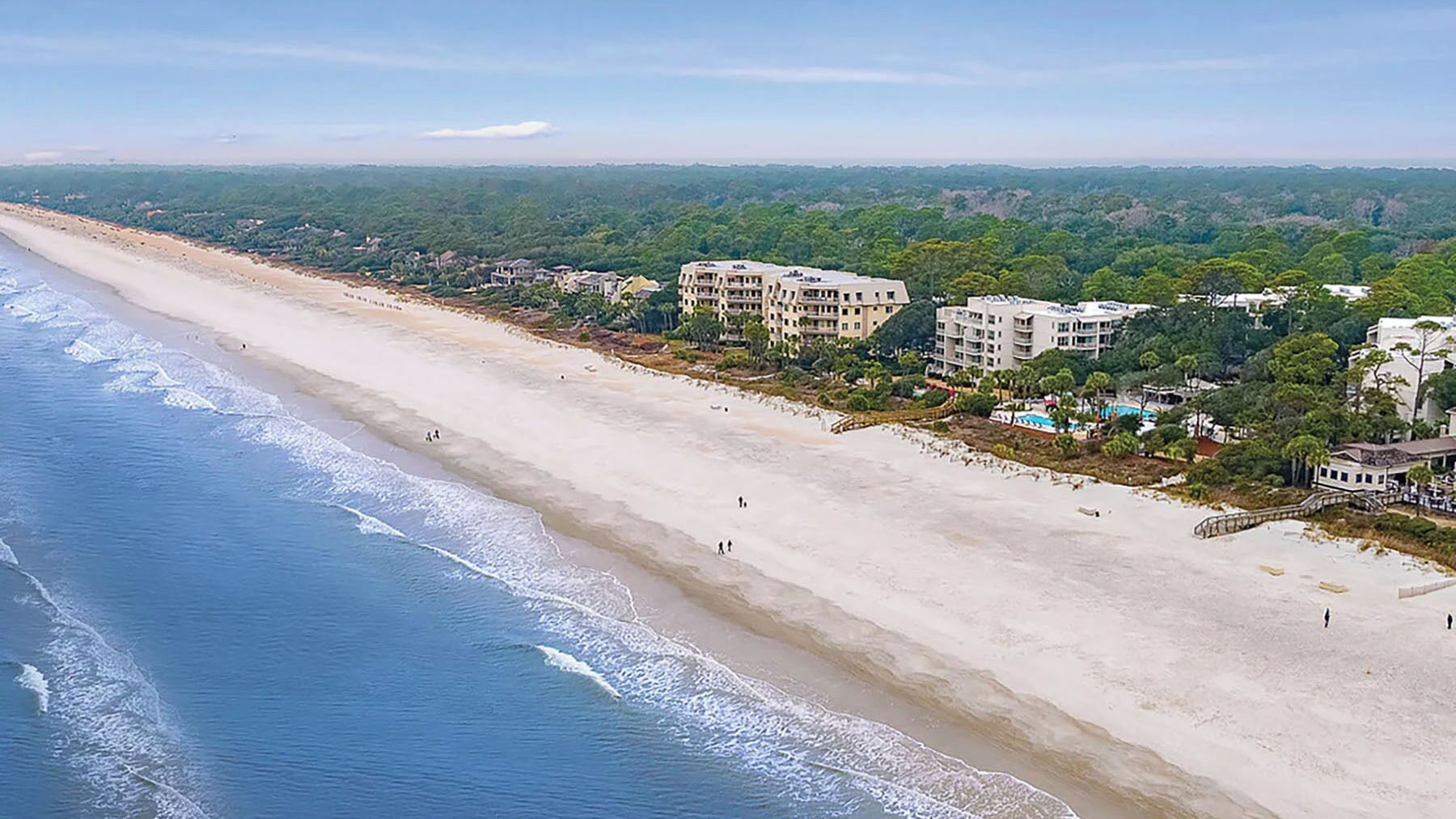 A coastal beach with a long stretch of sand, a few people walking, and several multi-story buildings in the background.