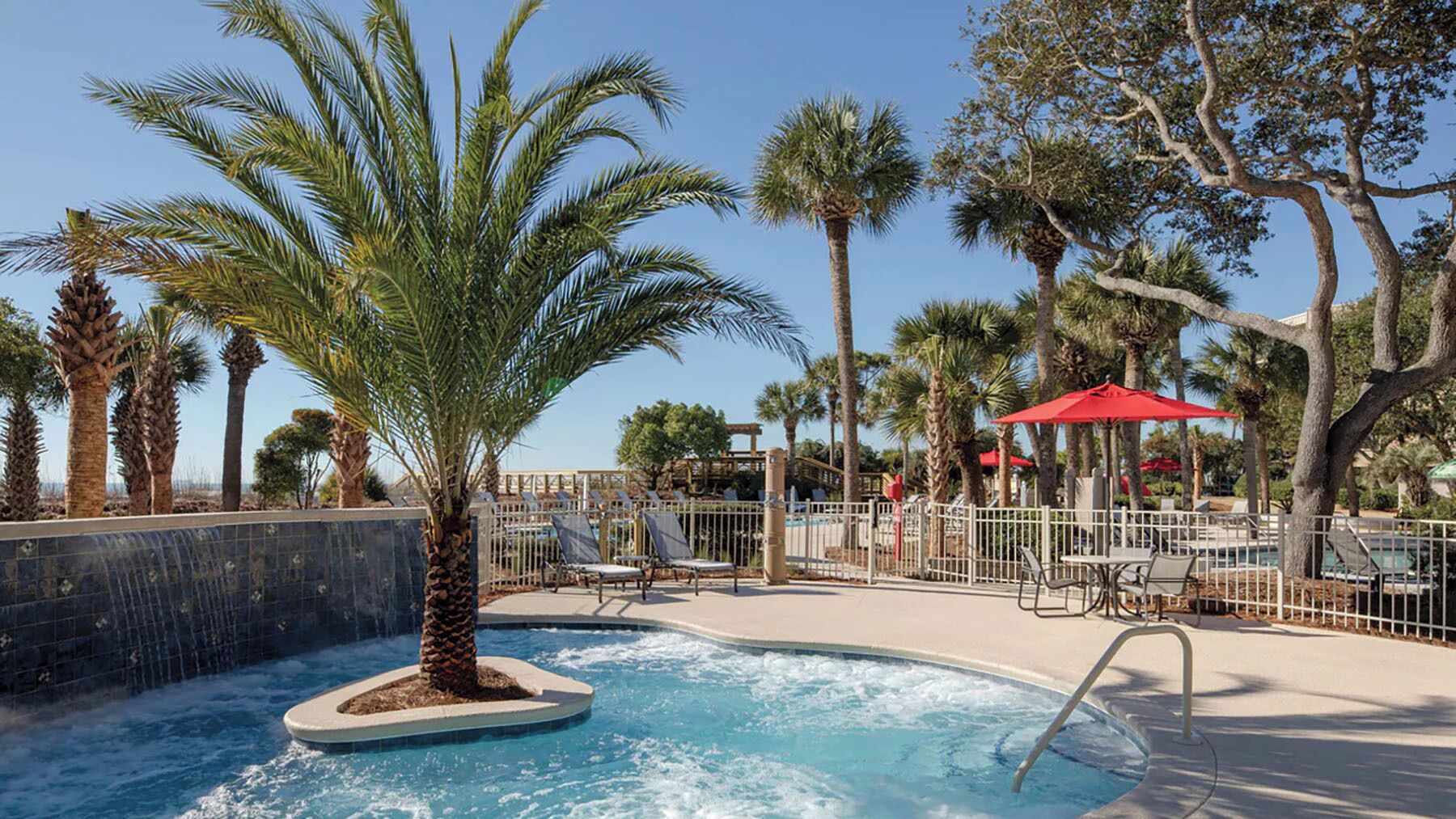 A pool area with palm trees, lounge chairs, and a red umbrella.