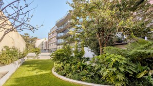 Área de jardim bem cuidada com um caminho curvo, cercada por vegetação exuberante e um edifício de vários andares com sacadas.