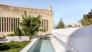 Piscina com deck revestido de azulejos, cercada por um muro branco baixo e um edifício com telhado e janelas em azulejos.
