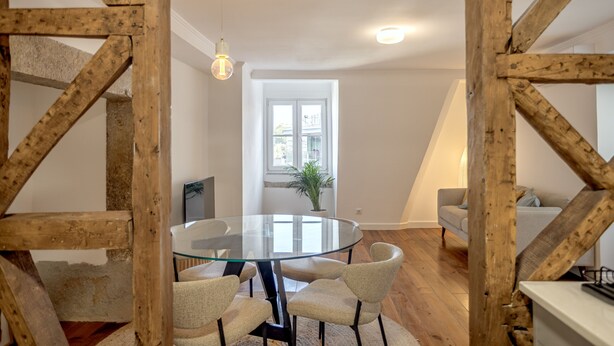 Uma sala de jantar com mesa de vidro cercada por cadeiras, vigas de madeira, um vaso com planta e uma janela com vista para edifícios.
